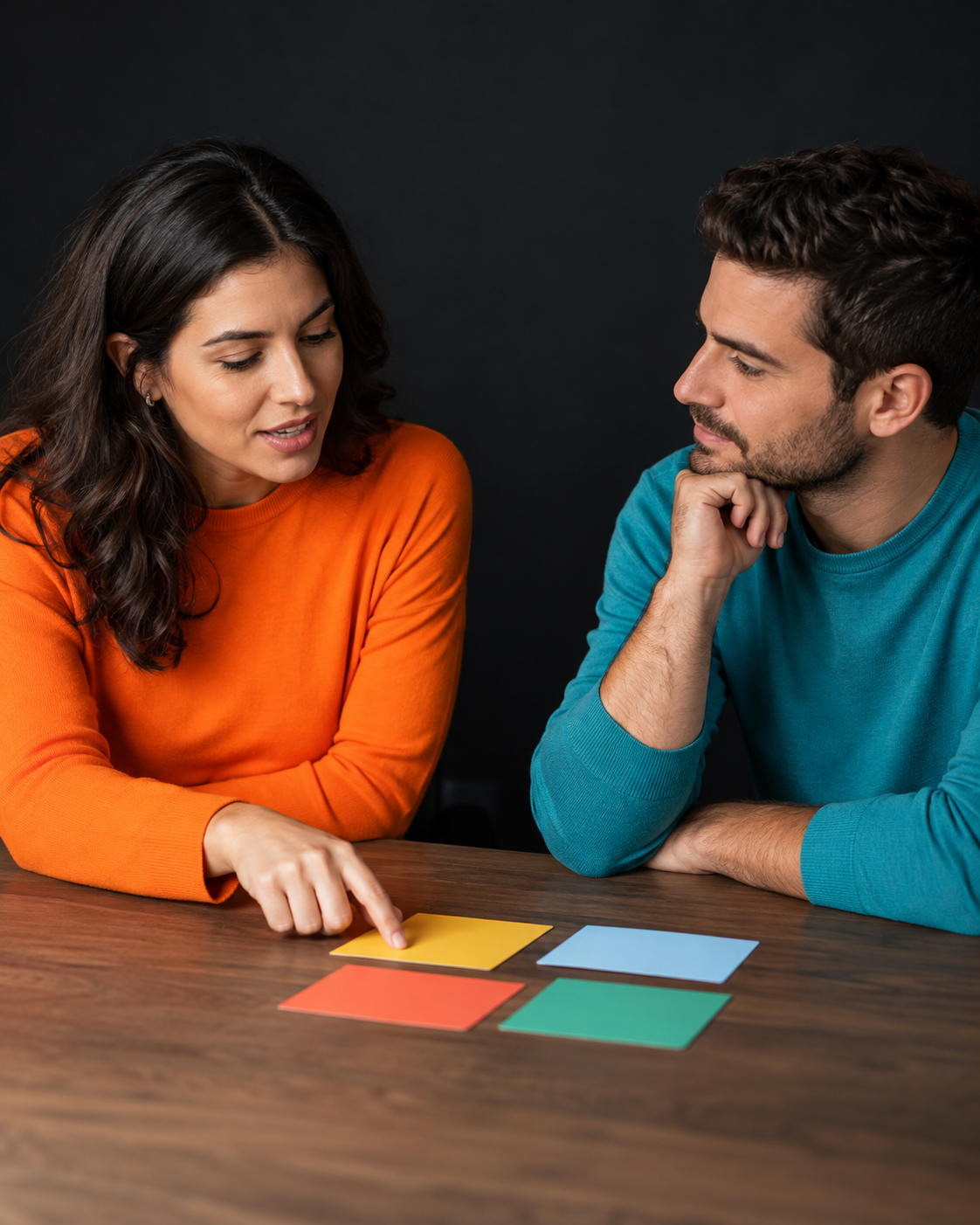 Duas pessoas adultas conversam diante de cartões sem texto, com roupa laranja e turquesa e fundo escuro.